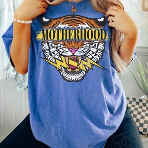May include: Blue t-shirt with a tiger graphic and the word "MOTHERHOOD". The tiger design features orange and black stripes, a pink nose, and a yellow lightning bolt. The shirt is a Flo Blue color.