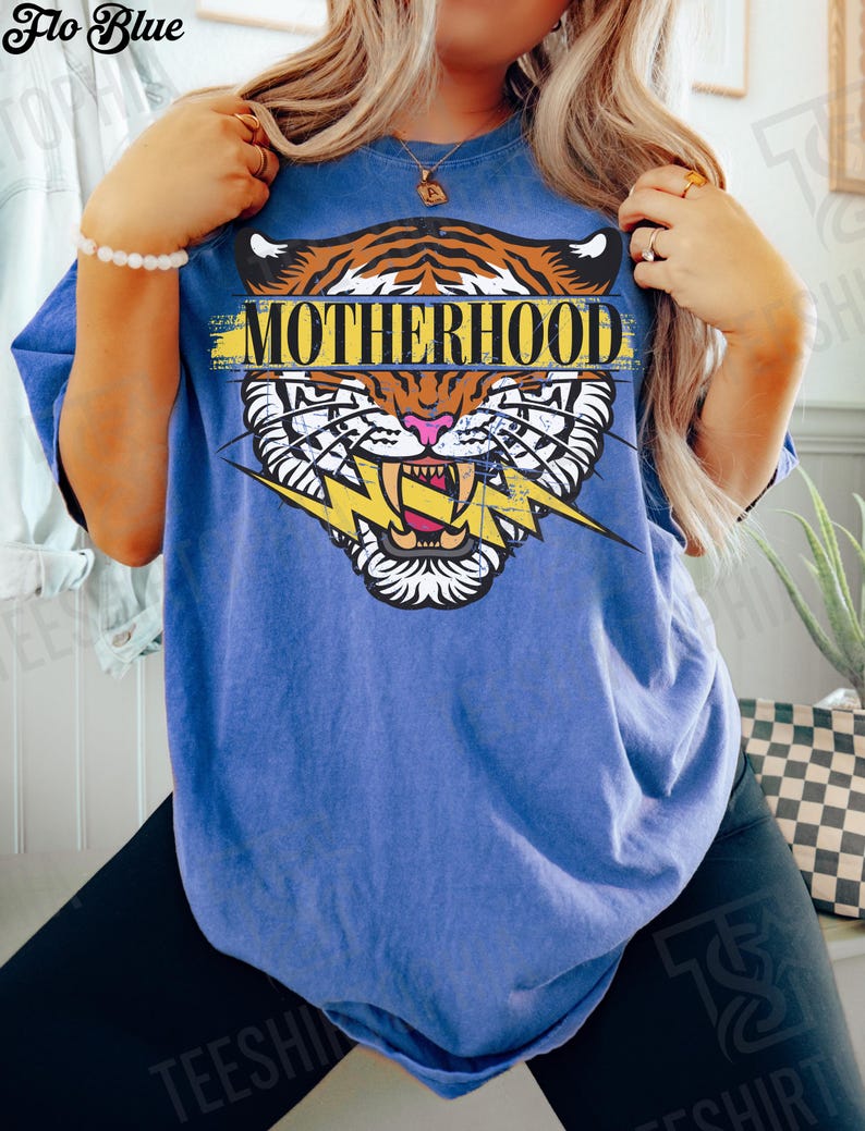 May include: Blue t-shirt with a tiger graphic and the word "MOTHERHOOD". The tiger design features orange and black stripes, a pink nose, and a yellow lightning bolt. The shirt is a Flo Blue color.