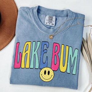 May include: A blue Comfort Colors t-shirt with the words "LAKE BUM" in colorful, retro-style lettering. A yellow smiley face with a checkered pattern is below the text. A gold necklace is draped across the shirt.
