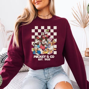 May include: A white ceramic mug with a black handle and the text "But First, Coffee" in a bold, black font. The mug is set against a blurred background, suggesting a cozy setting.