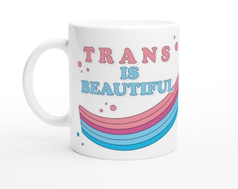 May include: White ceramic mug with a pink and blue rainbow design and the text "TRANS IS BEAUTIFUL" in pink and blue lettering.