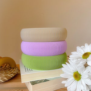 May include: Three stacked, ribbed, silicone rings in beige, lavender, and green. The rings are sitting on top of two books with a bouquet of white daisies in the background.