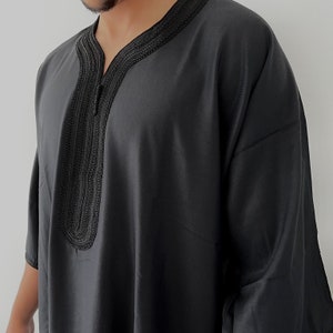 May include: A dark gray, short-sleeved garment with a V-neckline. The neckline and upper chest area are adorned with intricate black embroidery. The fabric appears smooth and drapes loosely. The garment has a relaxed fit.