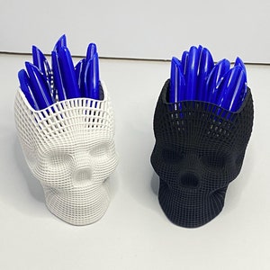 May include: Two 3D printed skull-shaped pencil holders, one white and one black, each filled with blue pens.