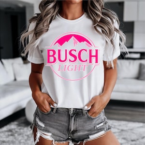 May include: White t-shirt featuring a pink graphic of the Busch Light logo. The design includes the brand name and a mountain range within a circular frame. The shirt has short sleeves and a crew neck.