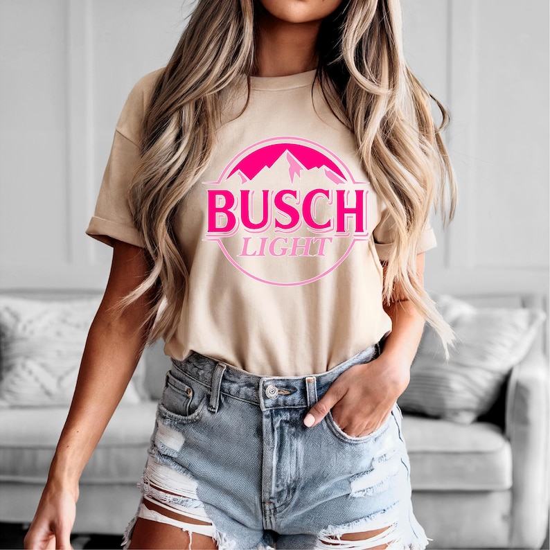 May include: Tan short-sleeved t-shirt featuring the "Busch Light" logo in pink. The logo includes a mountain range graphic inside a pink circle. The shirt is paired with denim shorts.