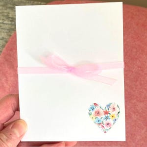May include: A white card tied with a sheer pink ribbon. A floral heart sticker with pink, blue, and yellow flowers is in the lower right corner. The card is held against a pink background.