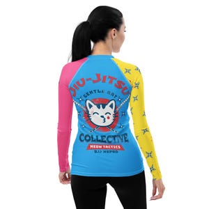 Könnte beinhalten: Ein blauer, rosa und gelber Rashguard mit langen Ärmeln, einem weißen Katzenmotiv und dem Text "Jiu-Jitsu Gentle Art Collective Meow Tactics BJJ NSPR D".