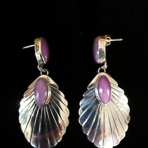 May include: A pair of silver drop earrings featuring a fan-shaped metal design with vertical ridges. Each earring is accented with two smooth, oval-shaped purple stones, one set at the top post and a larger one centred on the fan-shaped base.