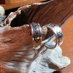 Op de afbeelding: Een paar zilveren oorringen met een breed, plat bandontwerp met gegraveerde, zwart ingevulde symbolen en patronen. De oorbellen hebben een gepolijste metalen afwerking en een sluiting met een steker, met gedetailleerde versieringen op het oppervlak.