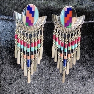 May include: A pair of silver-toned earrings featuring an oval inlay with a geometric pattern of blue, red, purple, and green segments. Below the oval, multiple dangling strands consist of small silver beads, red, blue, and black cylindrical beads.