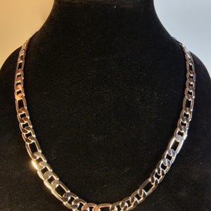 Peut inclure: Un collier chaîne de type Figaro argenté présentant un motif répétitif composé de trois petits maillons circulaires suivis d'un maillon ovale allongé. La surface métallique est polie et réfléchissante, créant des reflets sur les maillons.