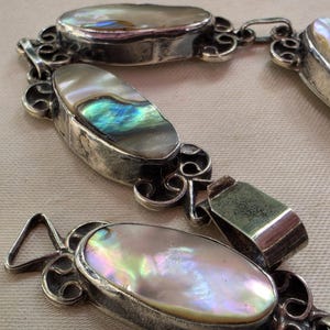 May include: A bracelet featuring oval-shaped iridescent abalone shell inlays set in tarnished silver-toned metal frames. The links are connected by decorative scrollwork metal components, showcasing a mix of pearlescent white, blue, and green hues.