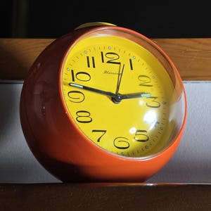 May include: A spherical alarm clock with a bright orange outer casing and a vibrant yellow circular face. The clock features black Arabic numerals, black hour and minute hands, a thin second hand, and the word Blessing printed in black.