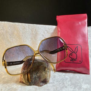 May include: Oversized sunglasses with amber-toned frames and gradient lenses, featuring the Playboy logo on the temple. Accompanied by a rectangular red storage pouch with a black Playboy rabbit logo printed on the front surface.