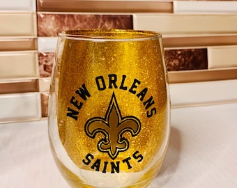 May include: A stemless wine glass with a gold glitter design. The glass features the words "New Orleans Saints" in black lettering, with the team's fleur-de-lis logo in the centre.