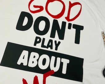 May include: White t-shirt with the text "GOD DON'T PLAY ABOUT ME" in red and black block letters. The words "GOD" and "ME" are in red, while "DON'T PLAY ABOUT" is in black.