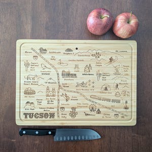 May include: A bamboo cutting board with a laser-etched map of Tucson, Arizona, featuring landmarks and place names. Two red apples rest above the board, and a black-handled knife is below. The board measures approximately 12 inches by 16 inches.