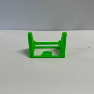 May include: A bright green plastic stand with a rectangular base and two upright sides. The stand is designed to hold a small object, such as a phone or a tablet.