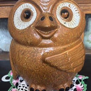 May include: A brown ceramic owl-shaped cookie jar with a lid. The owl has large white-rimmed eyes, a smiling face, and wings. It sits on a white and pink crocheted doily with green trim. The jar is a vintage kitchen item.