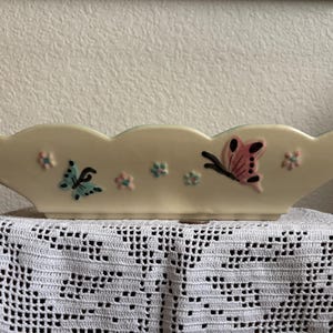 May include: Cream ceramic planter with a scalloped edge, decorated with blue and pink butterflies and small pink flowers. A decorative piece for displaying plants or flowers.