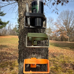 May include: Three Tree Caddy cup holders mounted on a tree trunk. The top holder is black and has the Tree Caddy logo. The middle holder is green and the bottom holder is orange.