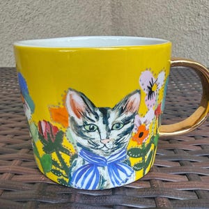 May include: Yellow ceramic mug with a gold handle and rim. The mug features a hand-painted design of a cat wearing a blue striped bow tie, surrounded by colorful flowers and foliage.