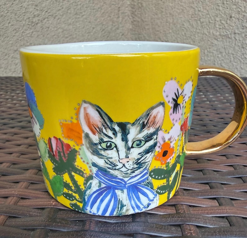 May include: Yellow ceramic mug with a gold handle and rim. The mug features a hand-painted design of a cat wearing a blue striped bow tie, surrounded by colorful flowers and foliage.