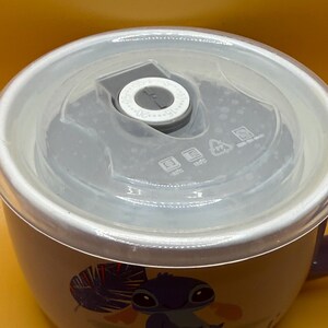 May include: A ceramic bowl with a clear lid. The bowl features a cartoon character illustration and the word "CHILL" in white. The lid has a gray center with a dial and measurement markings. The bowl is white with a purple handle.