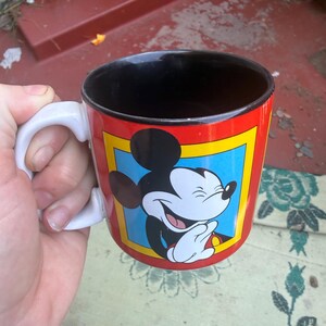 May include: A ceramic mug with a white handle and a black interior. The exterior features a red background with a yellow-bordered blue square containing a graphic illustration of Mickey Mouse laughing with his hands clasped together.
