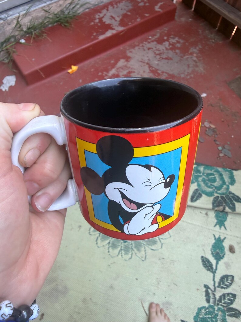 May include: A ceramic mug with a white handle and a black interior. The exterior features a red background with a yellow-bordered blue square containing a graphic illustration of Mickey Mouse laughing with his hands clasped together.