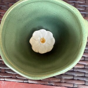 May include: A green ceramic bowl with a white flower-shaped accent in the center. The bowl has a rounded shape and a slightly textured surface. The flower has a yellow center.