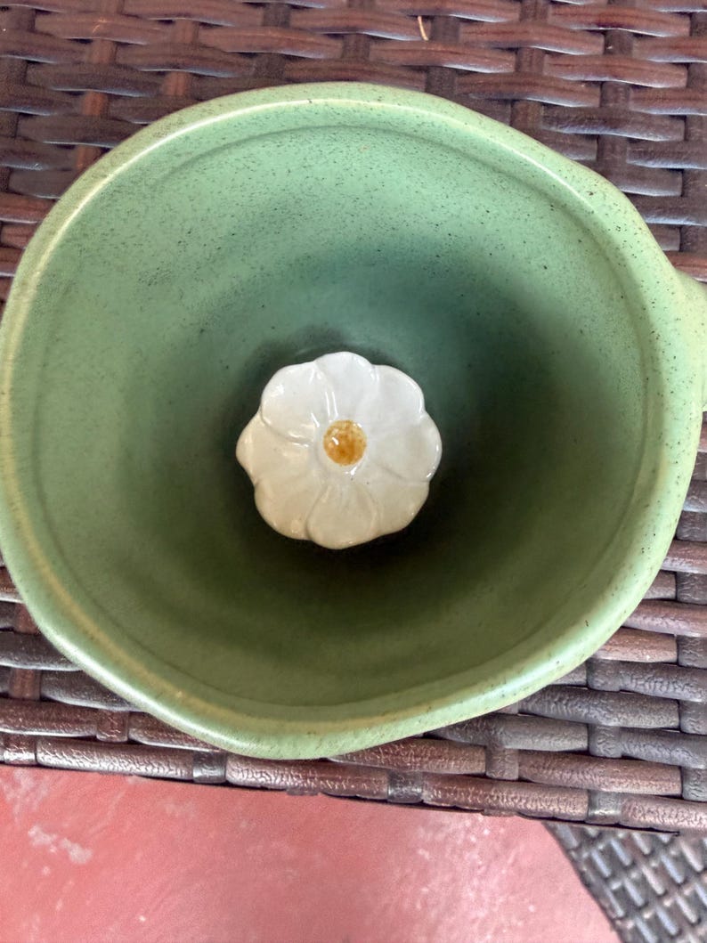 May include: A green ceramic bowl with a white flower-shaped accent in the center. The bowl has a rounded shape and a slightly textured surface. The flower has a yellow center.