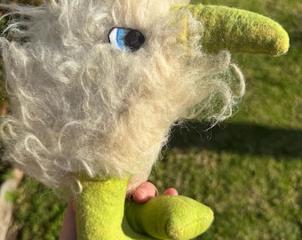 May include: A handmade, whimsical plush toy in the shape of a bird-like creature. It features a fluffy, off-white head with a blue and black eye, a long, green beak, and lime green feet. The toy is made of soft, textured materials.