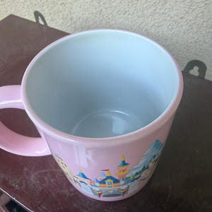 May include: A light pink ceramic mug with a white interior and a handle on the left side. The exterior features a colorful graphic illustration of a castle with blue, yellow, and red turrets, set against a light pink background.