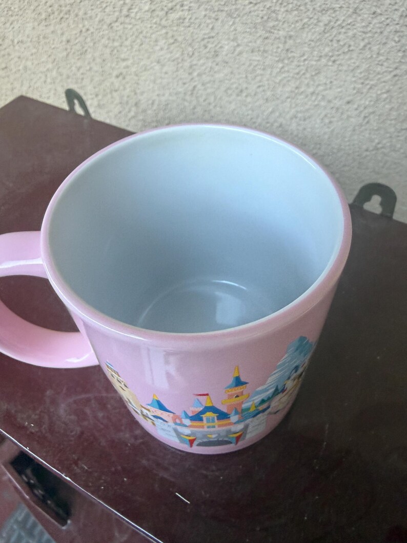 May include: A light pink ceramic mug with a white interior and a handle on the left side. The exterior features a colorful graphic illustration of a castle with blue, yellow, and red turrets, set against a light pink background.