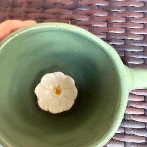 May include: A green ceramic bowl with a handle, featuring a white flower-shaped accent in the center. The flower has a yellow center. The bowl is held by a person.