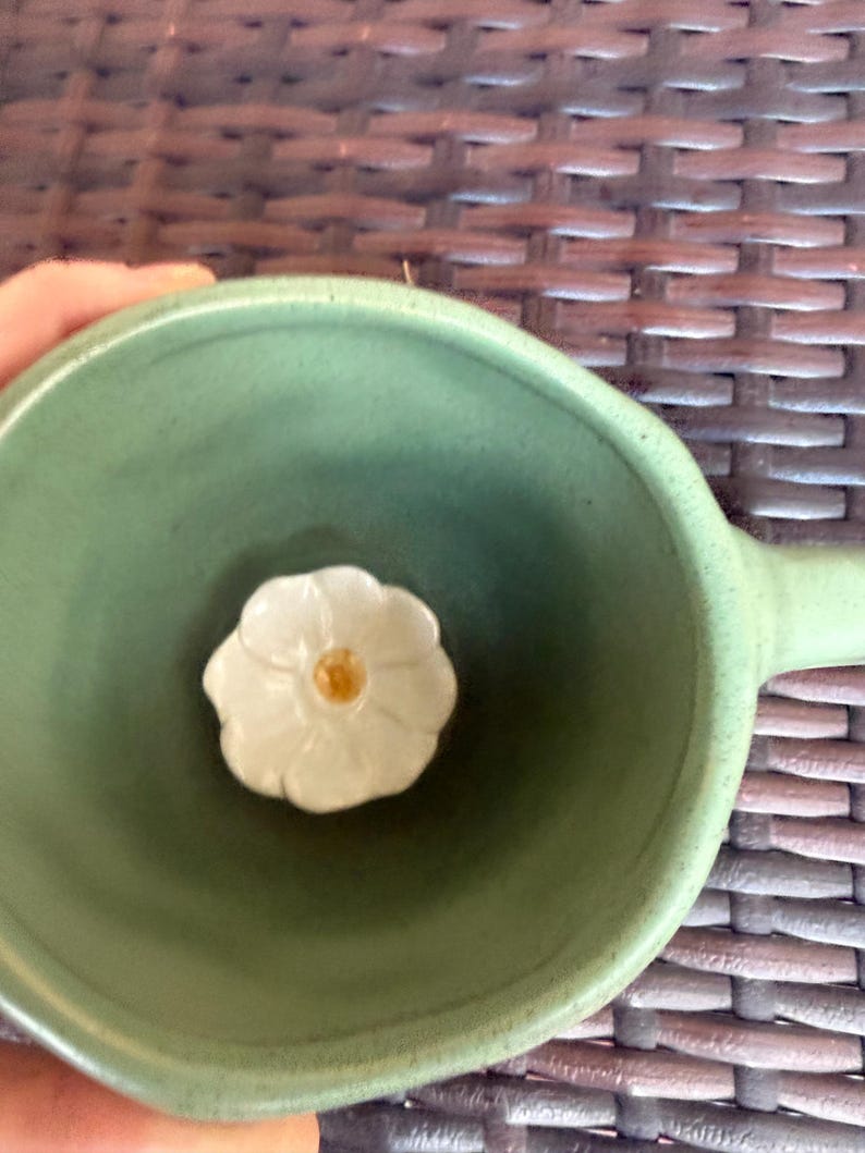 May include: A green ceramic bowl with a handle, featuring a white flower-shaped accent in the center. The flower has a yellow center. The bowl is held by a person.