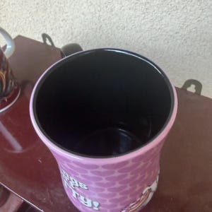 Puede incluir: Una taza de cer&aacute;mica con interior negro y exterior morado claro con un patr&oacute;n de corazones repetidos. La taza muestra texto blanco parcial que dice "gs" y "ry!" junto con un gr&aacute;fico de una porci&oacute;n de pizza en el lateral.