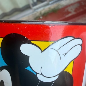 May include: A ceramic mug featuring a graphic of a cartoon character with a black head, white face, and white gloved hand raised against a vibrant red, yellow, and blue background. The rim of the mug is finished with a dark, glossy black border.