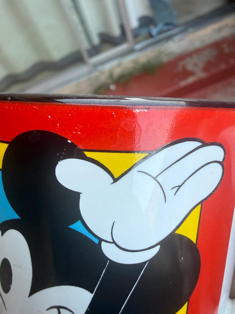 May include: A ceramic mug featuring a graphic of a cartoon character with a black head, white face, and white gloved hand raised against a vibrant red, yellow, and blue background. The rim of the mug is finished with a dark, glossy black border.