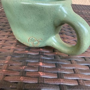 May include: A light green ceramic mug with a large handle and a slightly textured surface. The mug has a wide opening and a small decorative design near the base. The mug is sitting on a brown woven surface.