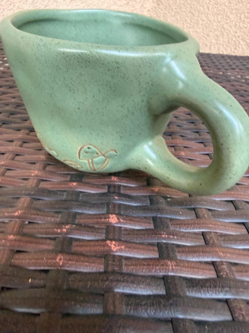 May include: A light green ceramic mug with a large handle and a slightly textured surface. The mug has a wide opening and a small decorative design near the base. The mug is sitting on a brown woven surface.