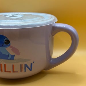 May include: A lavender ceramic mug with a white lid. The mug features a cartoon image of Stitch from Lilo & Stitch, with the word "ILLIN'" in blue and orange. The mug has a curved handle.
