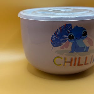 May include: A light purple bowl with a clear lid. The bowl features a cartoon character illustration and the word "CHILLI" in colorful letters. The bowl is likely used for food storage or serving.