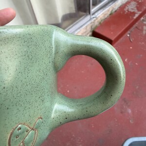 May include: A speckled, light green ceramic mug with a large handle. The mug features an embossed mushroom design. The mug is held by a hand, with the handle facing the viewer.