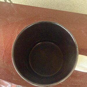 May include: A top-down view of a cylindrical mug featuring a dark brown interior and a white exterior handle. The mug has a smooth, solid-colored finish on the inside and a simple, functional design with a circular opening and a curved handle.