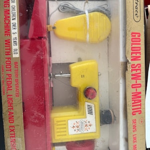 May include: A yellow toy sewing machine with a foot pedal, light, and extension table. The machine is battery operated and says "Straco Golden Sew-O-Matic Sews Like Mother's" on the box.