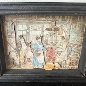May include: A framed, three-dimensional artwork depicting a vintage shop scene. The artwork features figures in period clothing, a scale, and various shop items. The frame is black, and the artwork is in muted colors.