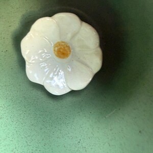 May include: A white ceramic flower-shaped lid with a yellow center. The lid is set against a green background, creating a contrast. The flower design adds a touch of elegance.